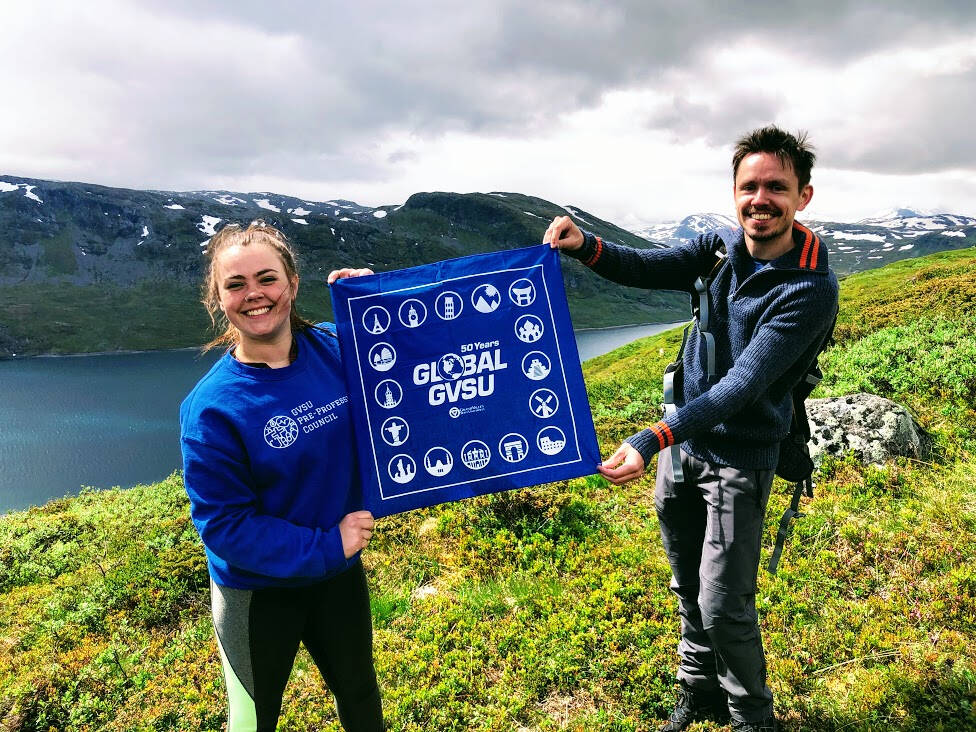 GVSU Students holding a Global GVSU sign on a green hill in Norway. The background shows cloudy skies and mountains.